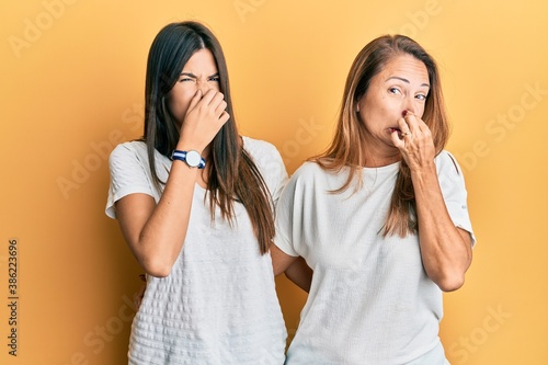 Hispanic family of mother and daughter wearing casual white tshirt smelling something stinky and disgusting, intolerable smell, holding breath with fingers on nose. bad smell