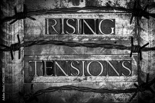 Rising Tensions text formed with real authentic typeset letters surrounded by barbed wire on vintage textured monochrome background  © IHX