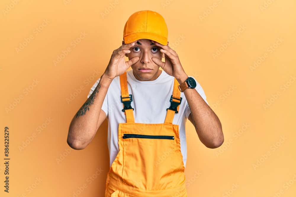 Young handsome african american man wearing handyman uniform over yellow background trying to open eyes with fingers, sleepy and tired for morning fatigue
