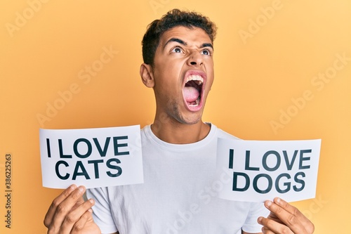 Young handsome african american man holding paper with i love cats and i love dogs phrase angry and mad screaming frustrated and furious, shouting with anger looking up.