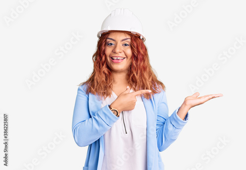 Young latin woman wearing architect hardhat amazed and smiling to the camera while presenting with hand and pointing with finger.