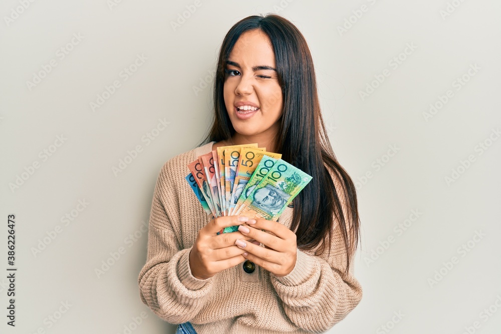 Young beautiful hispanic girl holding australian dollars winking looking at the camera with sexy expression, cheerful and happy face. Young beautiful hispanic girl holding australian dollars winking looking at the camera with sexy expression, cheerful and happy face.