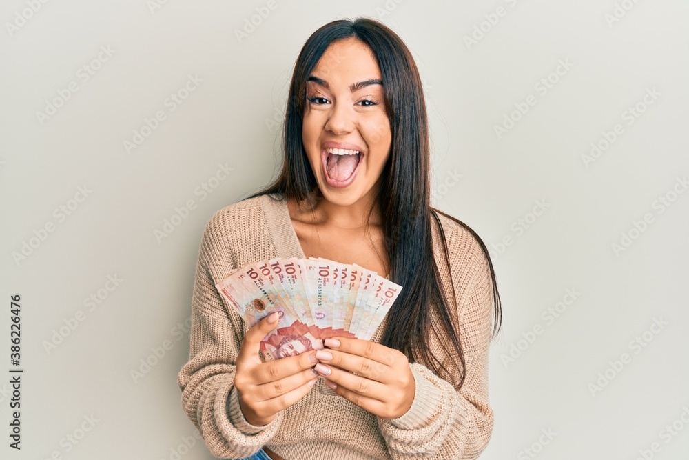 Young beautiful hispanic girl holding 10 colombian pesos banknotes celebrating crazy and amazed for success with open eyes screaming excited. Young beautiful hispanic girl holding 10 colombian pesos banknotes celebrating crazy and amazed for success with open eyes screaming excited.