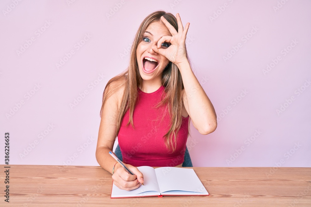 Young caucasian girl writing notebook sitting on the table smiling happy doing ok sign with hand on eye looking through fingers Young caucasian girl writing notebook sitting on the table smiling happy doing ok sign with hand on eye looking through fingers