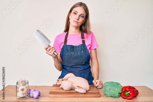 Young blonde girl wearing cook apron cooking chicken thinking attitude and sober expression looking self confident