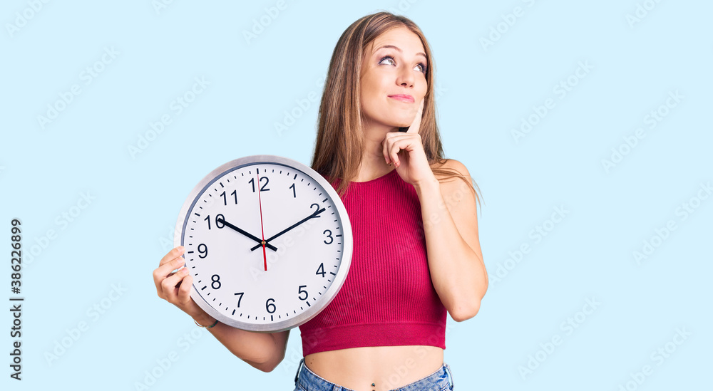Young beautiful blonde woman holding big clock serious face thinking about question with hand on chin, thoughtful about confusing idea Young beautiful blonde woman holding big clock serious face thinking about question with hand on chin, thoughtful about confusing idea