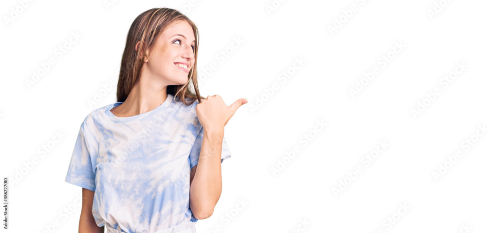 Young beautiful blonde woman wearing tye die tshirt smiling with happy face looking and pointing to the side with thumb up. Young beautiful blonde woman wearing tye die tshirt smiling with happy face looking and pointing to the side with thumb up.