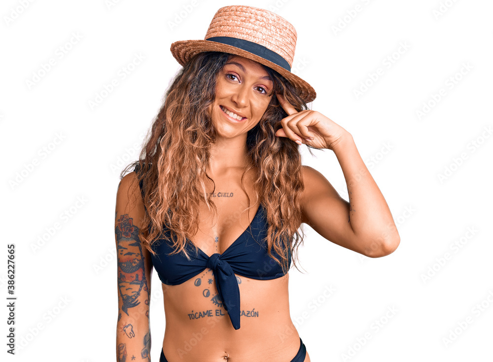 Young hispanic woman with tattoo wearing bikini and summer hat smiling pointing to head with one finger, great idea or thought, good memory Young hispanic woman with tattoo wearing bikini and summer hat smiling pointing to head with one finger, great idea or thought, good memory