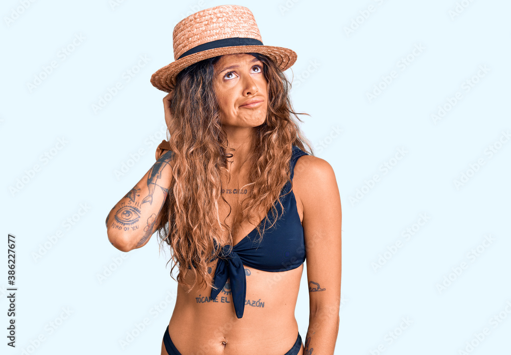 Young hispanic woman with tattoo wearing bikini and summer hat confuse and wondering about question. uncertain with doubt, thinking with hand on head. pensive concept. Young hispanic woman with tattoo wearing bikini and summer hat confuse and wondering about question. uncertain with doubt, thinking with hand on head. pensive concept.