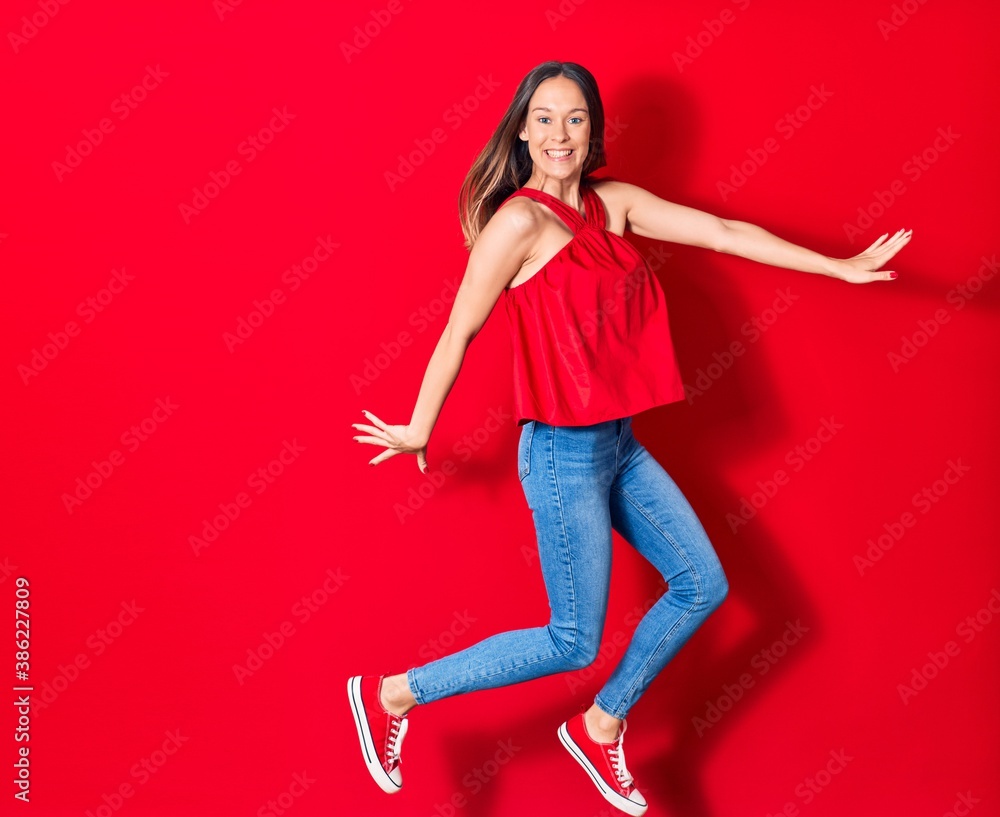 Young beautiful woman wearing casual clothes smiling happy. Jumping with smile on face and arms opened over isolated red background Young beautiful woman wearing casual clothes smiling happy. Jumping with smile on face and arms opened over isolated red background