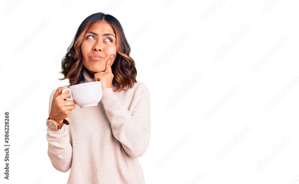 Young beautiful mixed race woman holding a cup of coffee serious face thinking about question with hand on chin, thoughtful about confusing idea Young beautiful mixed race woman holding a cup of coffee serious face thinking about question with hand on chin, thoughtful about confusing idea
