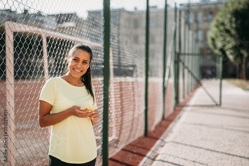 Smiling sporty woman standing near fence with smartphone