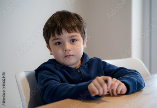 Portrait handsome boy looking at camera with smiling face, Positive child sitting alone waiting for food in dining room, Kid wearing hoodie looking out deep in thought