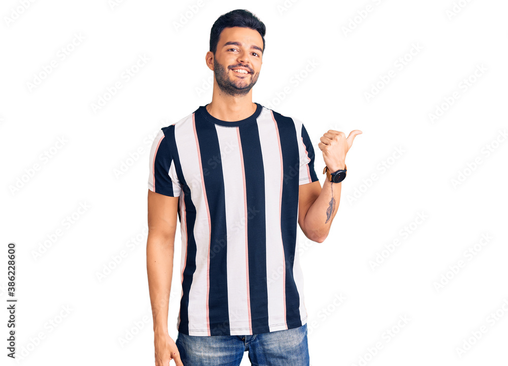 Young handsome hispanic man wearing striped tshirt smiling with happy face looking and pointing to the side with thumb up. Young handsome hispanic man wearing striped tshirt smiling with happy face looking and pointing to the side with thumb up.