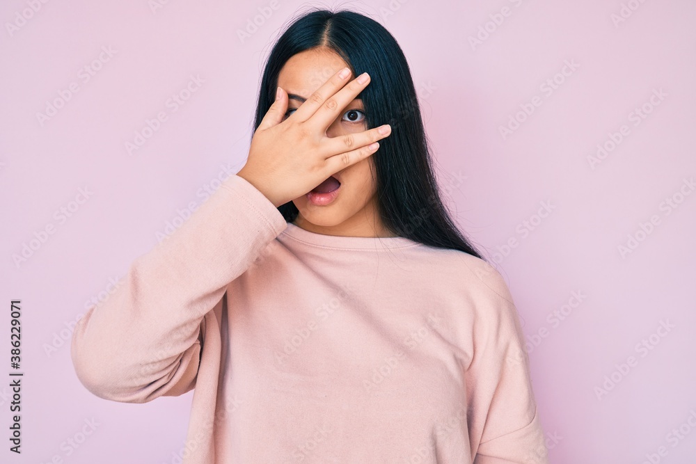 Young beautiful asian girl wearing casual pink sweater peeking in shock covering face and eyes with hand, looking through fingers afraid Young beautiful asian girl wearing casual pink sweater peeking in shock covering face and eyes with hand, looking through fingers afraid