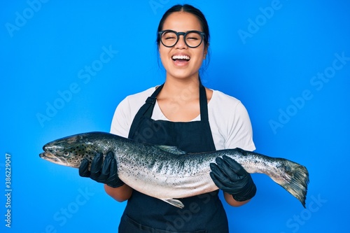 Young beautiful asian girl fishmonger selling fresh raw salmon smiling and laughing hard out loud because funny crazy joke.
