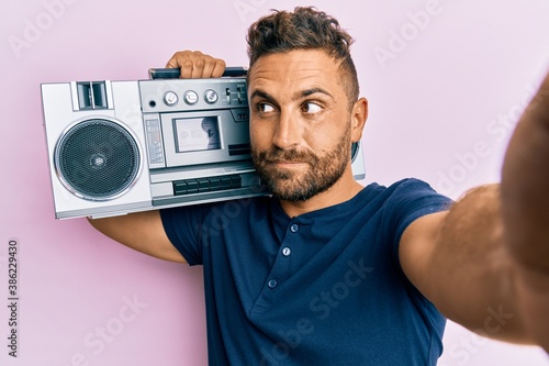 Handsome man with beard holding boombox, listening to music smiling looking to the side and staring away thinking.