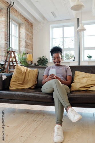 Restful young African woman with smartphone sitting on black leather couch