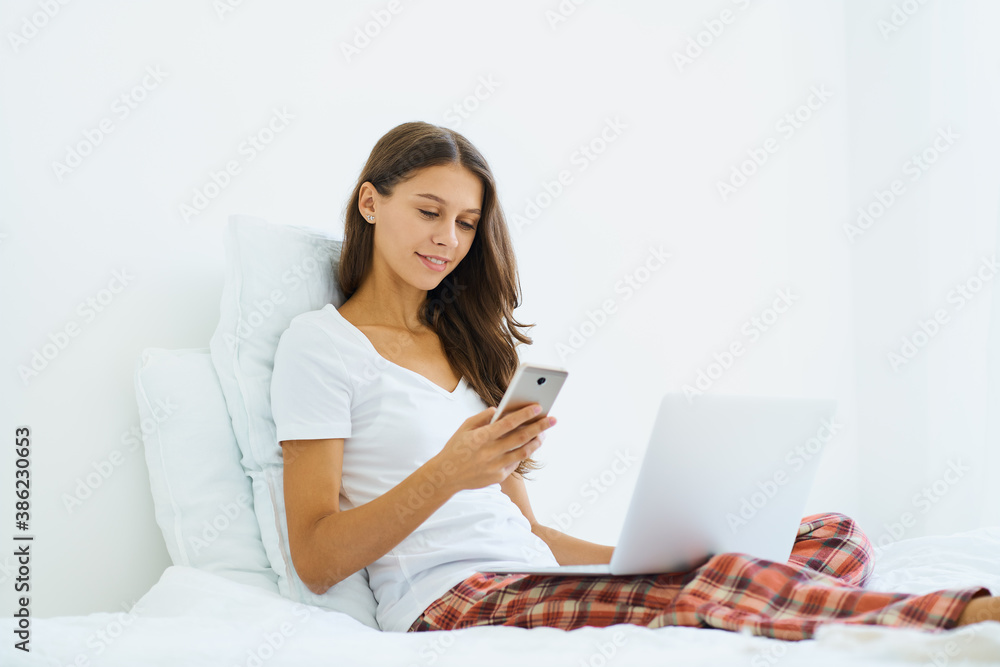 Young woman shopping online, entering code from SMS message on mobile phone, online shopping security. Female sitting on bed in bedroom and working from home on laptop Young woman shopping online, entering code from SMS message on mobile phone, online shopping security. Female sitting on bed in bedroom and working from home on laptop