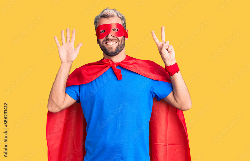 Young blond man wearing super hero custome showing and pointing up with fingers number seven while smiling confident and happy.