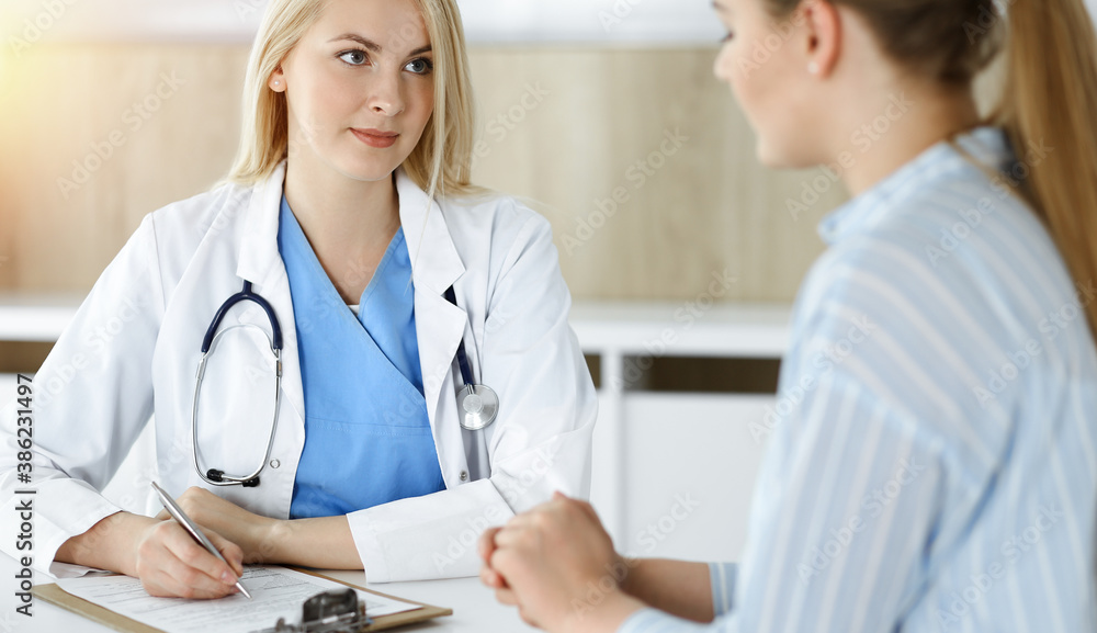 Woman-doctor and patient sitting and talking in sunny clinic. Blonde therapist is cheerfully smiling Woman-doctor and patient sitting and talking in sunny clinic. Blonde therapist is cheerfully smiling