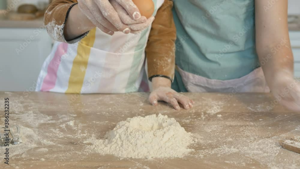 Happy little girl with young mother breaks egg into flour to make pie dough on large wooden table in light kitchen closeup Happy little girl with young mother breaks egg into flour to make pie dough on large wooden table in light kitchen closeup