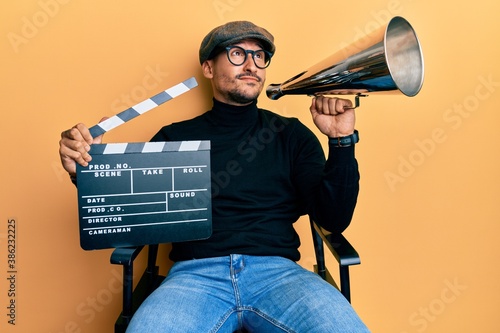 Картината върху платно Handsome man with tattoos holding video film clapboard and louder smiling looking to the side and staring away thinking