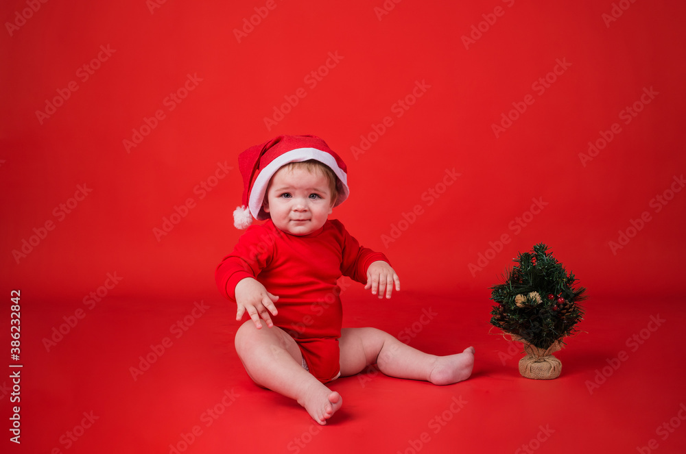 funny baby girl in a new year's hat and bodysuit sitting with a small Christmas tree on a red background with space for text funny baby girl in a new year's hat and bodysuit sitting with a small Christmas tree on a red background with space for text