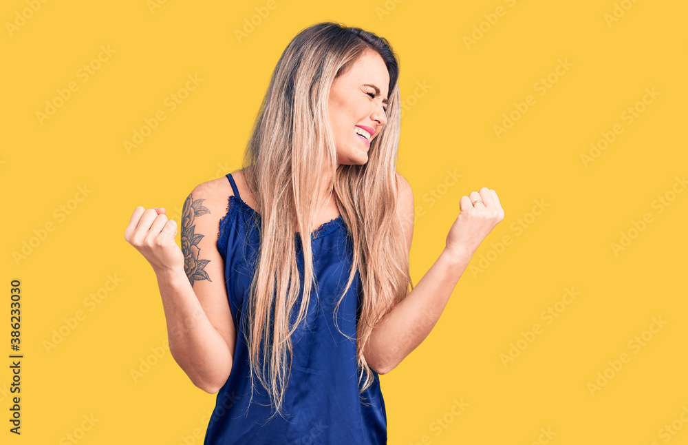 Young beautiful blonde woman wearing casual sleeveless t-shirt very happy and excited doing winner gesture with arms raised, smiling and screaming for success. celebration concept. Young beautiful blonde woman wearing casual sleeveless t-shirt very happy and excited doing winner gesture with arms raised, smiling and screaming for success. celebration concept.