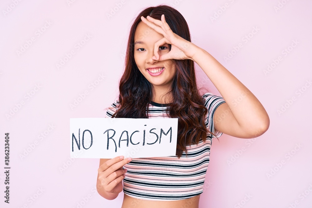 Young beautiful chinese girl holding no racism banner smiling happy doing ok sign with hand on eye looking through fingers Young beautiful chinese girl holding no racism banner smiling happy doing ok sign with hand on eye looking through fingers