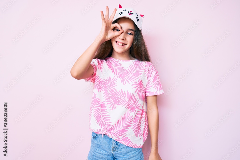Cute hispanic child girl wearing casual clothes and funny kitty cap smiling happy doing ok sign with hand on eye looking through fingers Cute hispanic child girl wearing casual clothes and funny kitty cap smiling happy doing ok sign with hand on eye looking through fingers