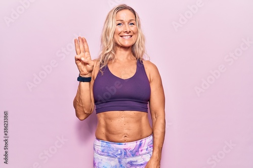 Middle age caucasian blonde woman wearing sportswear over pink background showing and pointing up with fingers number three while smiling confident and happy. © Krakenimages.com