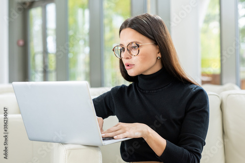 Serious young businesswoman in pullover and eyeglasses looking at laptop display