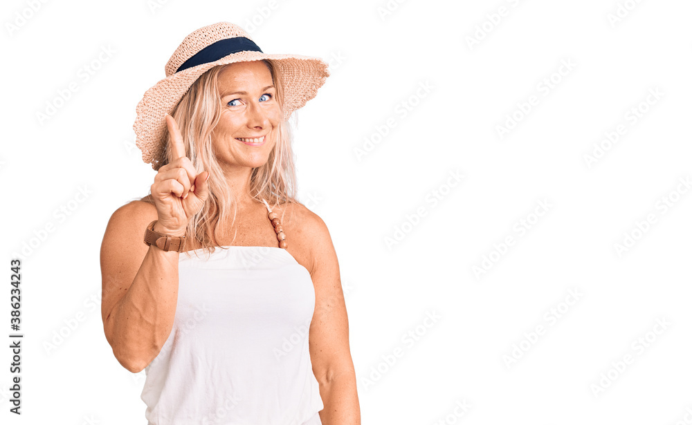 Middle age fit blonde woman wearing summer hat showing and pointing up with finger number one while smiling confident and happy. Middle age fit blonde woman wearing summer hat showing and pointing up with finger number one while smiling confident and happy.