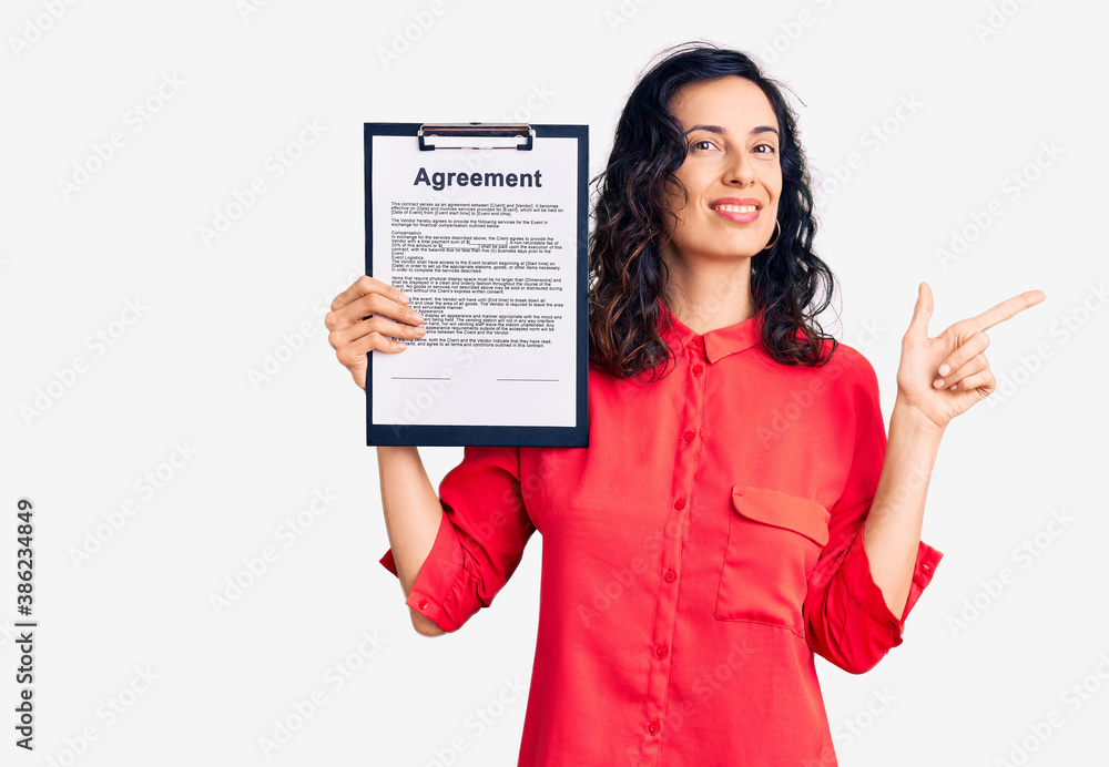 Young beautiful hispanic woman holding clipboard with agreement document smiling happy pointing with hand and finger to the side Young beautiful hispanic woman holding clipboard with agreement document smiling happy pointing with hand and finger to the side