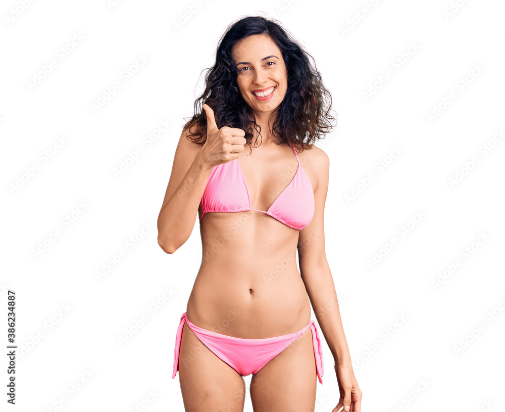 Young beautiful hispanic woman wearing bikini doing happy thumbs up gesture with hand. approving expression looking at the camera showing success. Young beautiful hispanic woman wearing bikini doing happy thumbs up gesture with hand. approving expression looking at the camera showing success.