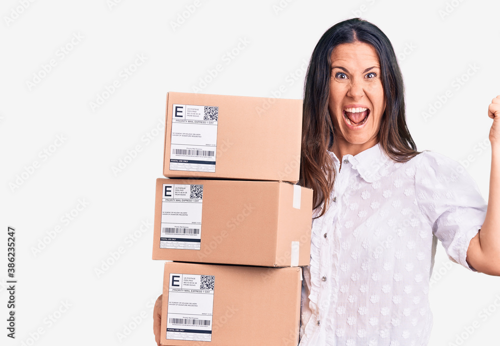 Young beautiful brunette woman holding delivery cardboard package screaming proud, celebrating victory and success very excited with raised arms Young beautiful brunette woman holding delivery cardboard package screaming proud, celebrating victory and success very excited with raised arms