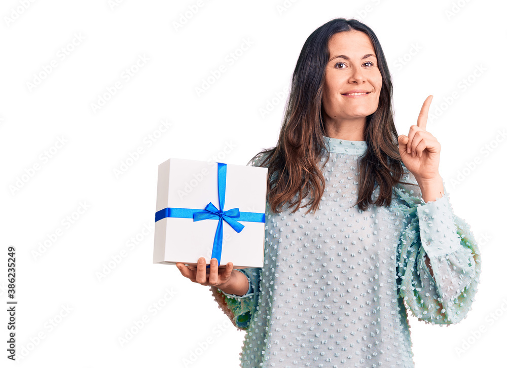 Young beautiful brunette woman holding birthday gift surprised with an idea or question pointing finger with happy face, number one Young beautiful brunette woman holding birthday gift surprised with an idea or question pointing finger with happy face, number one