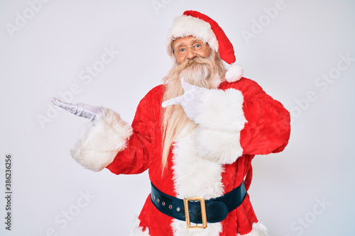 Old senior man with grey hair and long beard wearing santa claus costume with suspenders amazed and smiling to the camera while presenting with hand and pointing with finger.