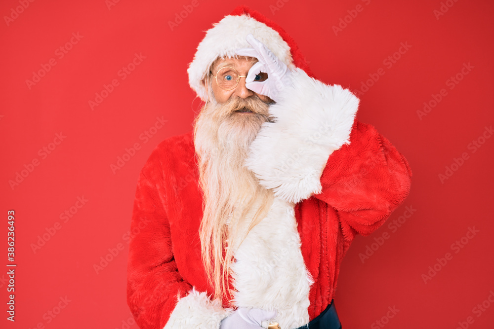 Old senior man with grey hair and long beard wearing traditional santa claus costume doing ok gesture with hand smiling, eye looking through fingers with happy face. Old senior man with grey hair and long beard wearing traditional santa claus costume doing ok gesture with hand smiling, eye looking through fingers with happy face.