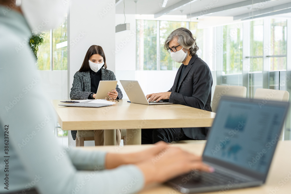 Two busy colleagues in smart casualwear and protective masks networking by table Two busy colleagues in smart casualwear and protective masks networking by table