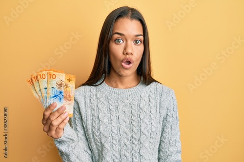 Beautiful hispanic woman holding 10 swiss franc banknotes scared and amazed with open mouth for surprise, disbelief face