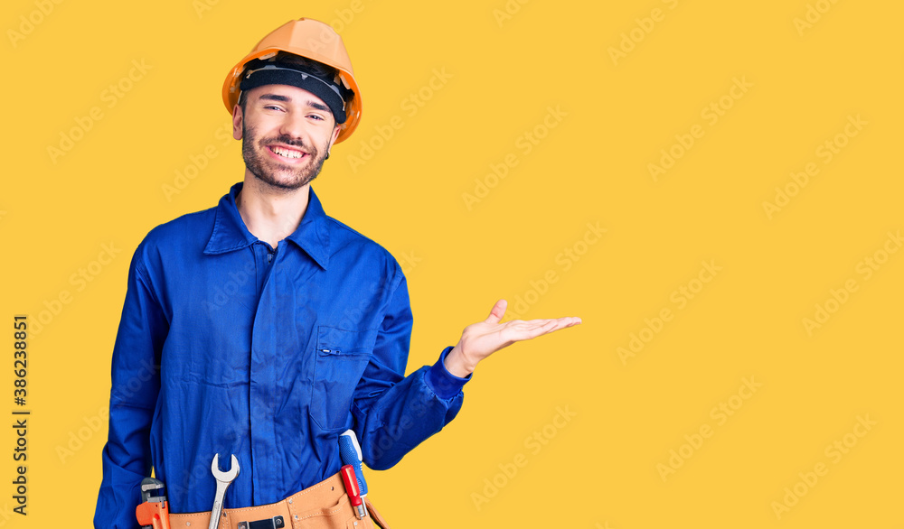 Young hispanic man wearing worker uniform smiling cheerful presenting and pointing with palm of hand looking at the camera. Young hispanic man wearing worker uniform smiling cheerful presenting and pointing with palm of hand looking at the camera.