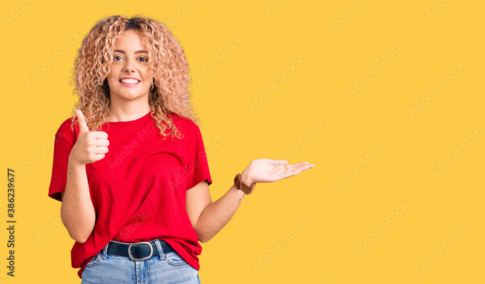Young blonde woman with curly hair wearing casual red tshirt showing palm hand and doing ok gesture with thumbs up, smiling happy and cheerful