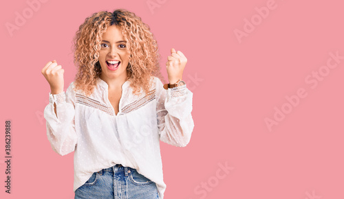 Young blonde woman with curly hair wearing elegant summer shirt celebrating surprised and amazed for success with arms raised and open eyes. winner concept.