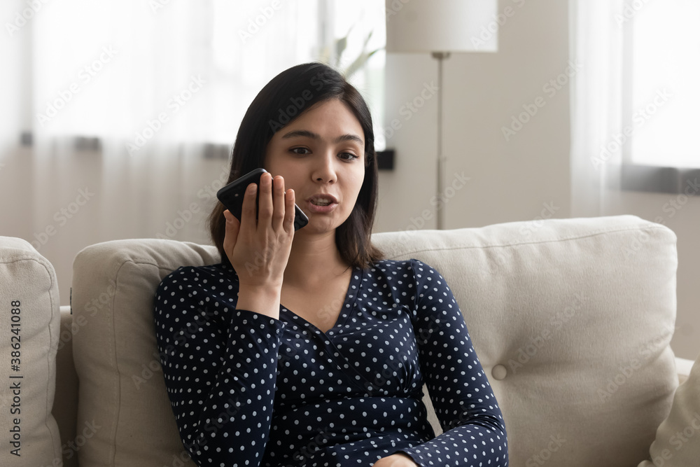 Young Vietnamese woman sit on sofa at home talk speak on loudspeaker on cellphone, activate voice recognition assistant app. Asian girl record audio message on smartphone. Technology concept. Young Vietnamese woman sit on sofa at home talk speak on loudspeaker on cellphone, activate voice recognition assistant app. Asian girl record audio message on smartphone. Technology concept.