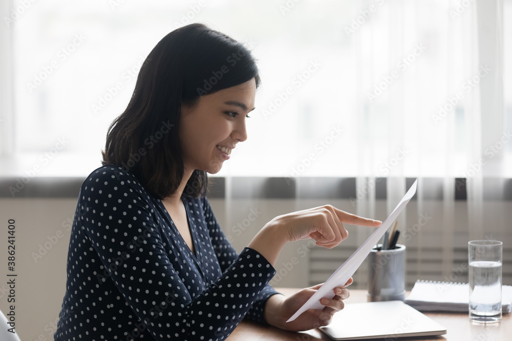 Happy young Vietnamese woman sit at desk at home office read good positive news in paper letter. Smiling millennial asian girl consider message or promotion offer notice in postal correspondence.