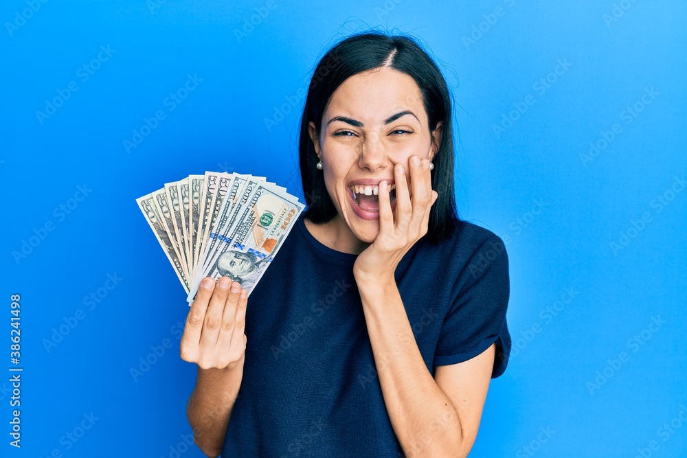 Beautiful young woman holding dollars laughing and embarrassed giggle covering mouth with hands, gossip and scandal concept Beautiful young woman holding dollars laughing and embarrassed giggle covering mouth with hands, gossip and scandal concept