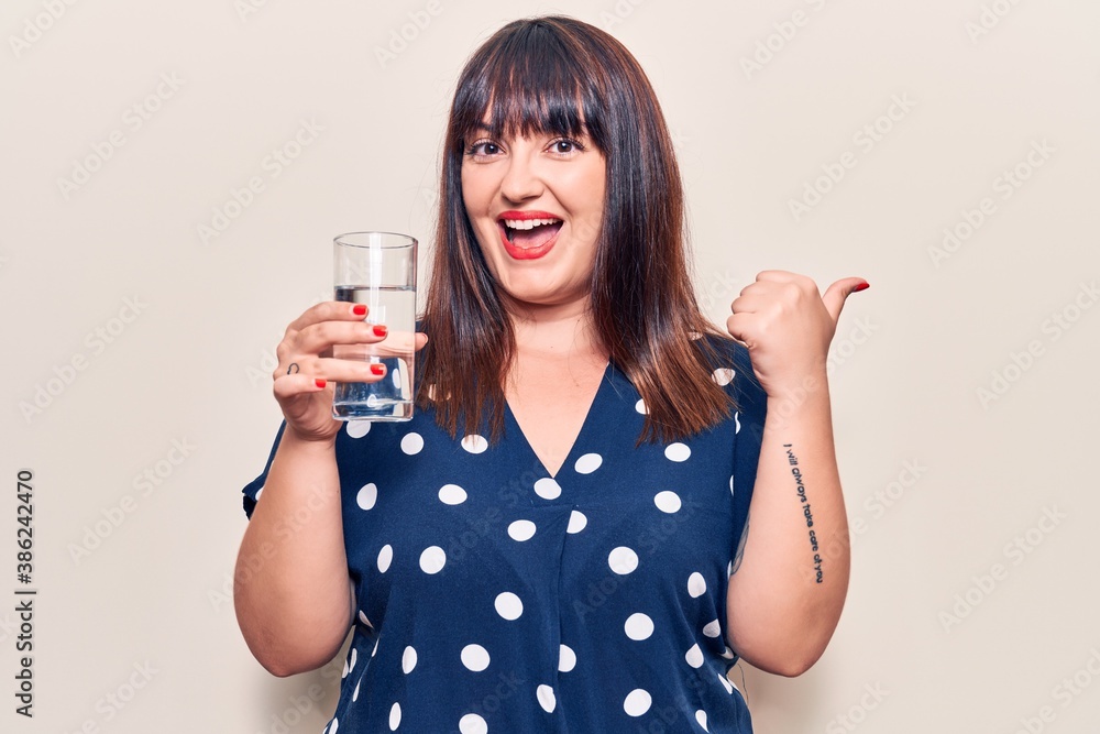 Young plus size woman drinking glass of water pointing thumb up to the side smiling happy with open mouth Young plus size woman drinking glass of water pointing thumb up to the side smiling happy with open mouth