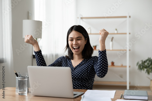 Excited young Vietnamese woman sit at desk at home office triumph reading good unexpected news or email on laptop. Overjoyed asian female look at computer screen feel euphoric win lottery online.